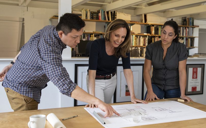 three light-skinned people examing a set of blueprints on a light wood table