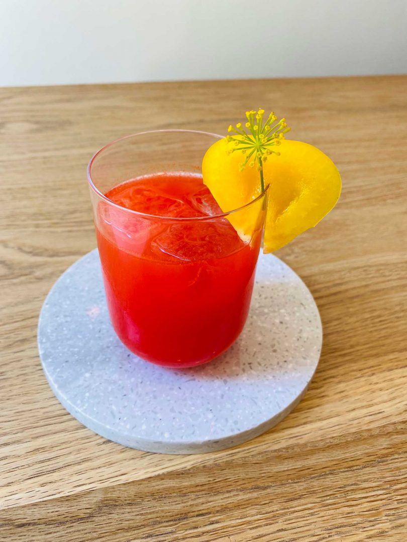 looking down at red cocktail in a clear drinking glass with yellow adornment sitting on white trivet on wood table