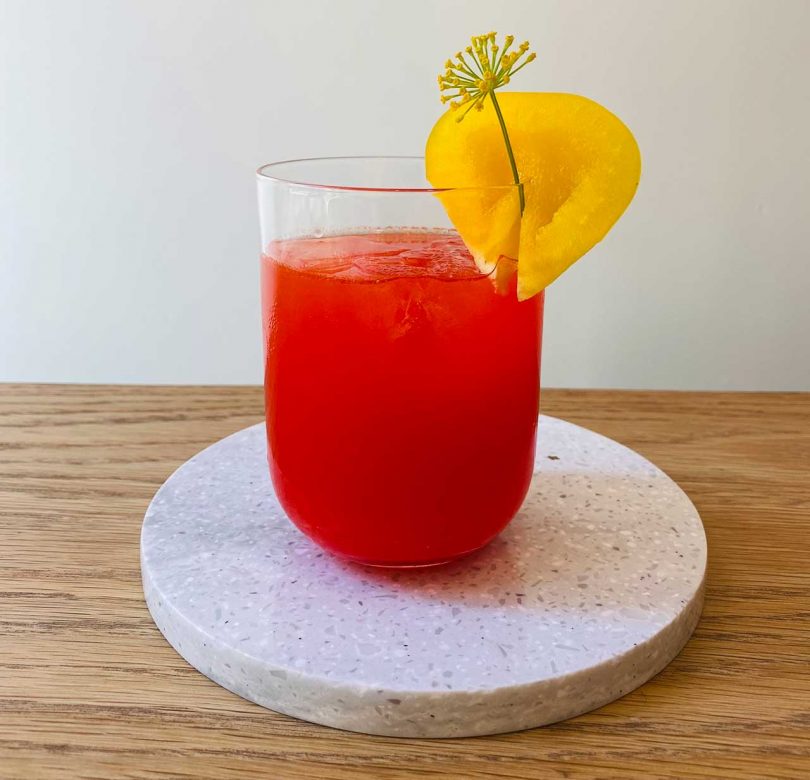 Red cocktail in a transparent glass with yellow decoration sitting on a white trivet on a wooden table