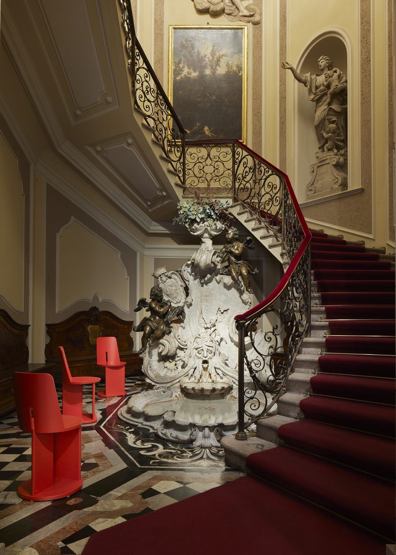 three modern vermillion stools gathered around a traditional water fountain at the base of an ornate curved staircase