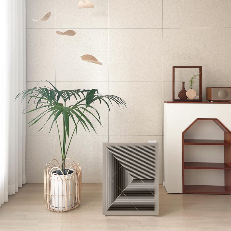 Airmega 240 on light oak wood floor with off-white panels wall, mobile hanging nearby with palm house plant and small decorative shelf to the right.