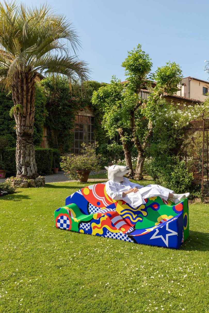 Outside in lush green landscape, a colorful, curvy sofa lying with person on top