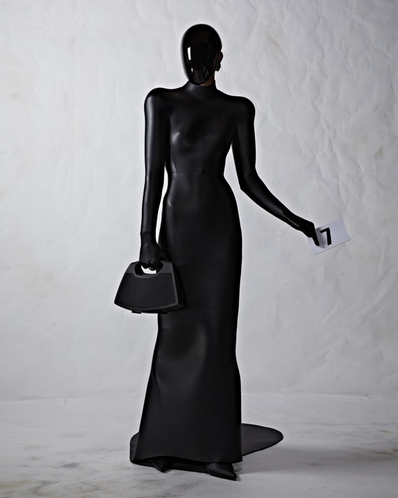 Balenciaga couture model in black floor length rubber dress and in full face mask holding black Bang & Olufsen Speaker Bag.