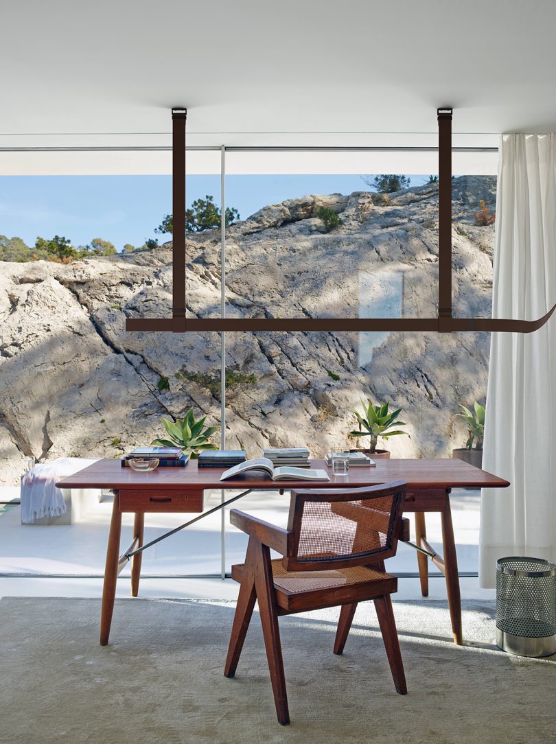 belt-like light fixture hanging over a desk and chair with a landscape view through the window