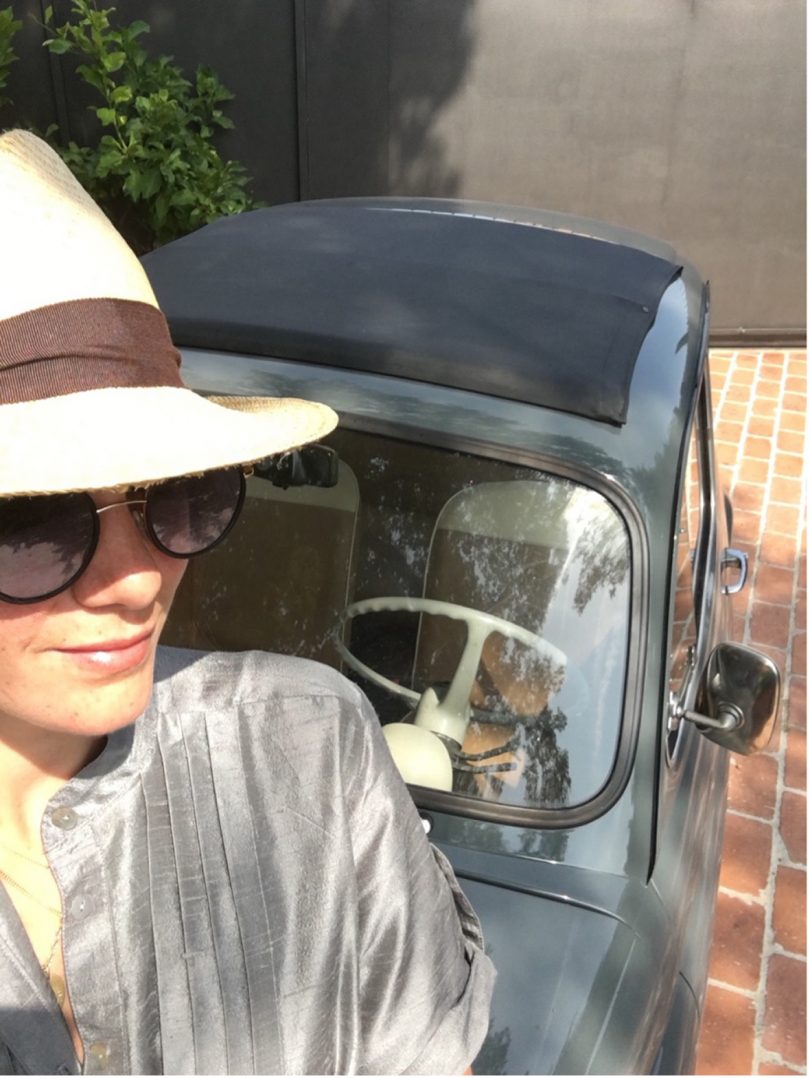 light-skinned woman wearing a brimmed hat and sunglasses taking a selfie of herself and a dark car