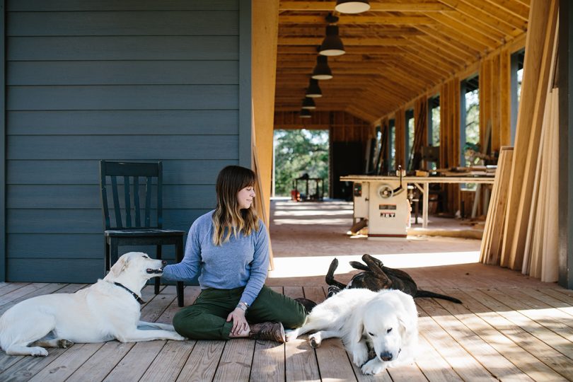 light-skinned woman with long, dark hair sits on an outdoor deck with three dogs