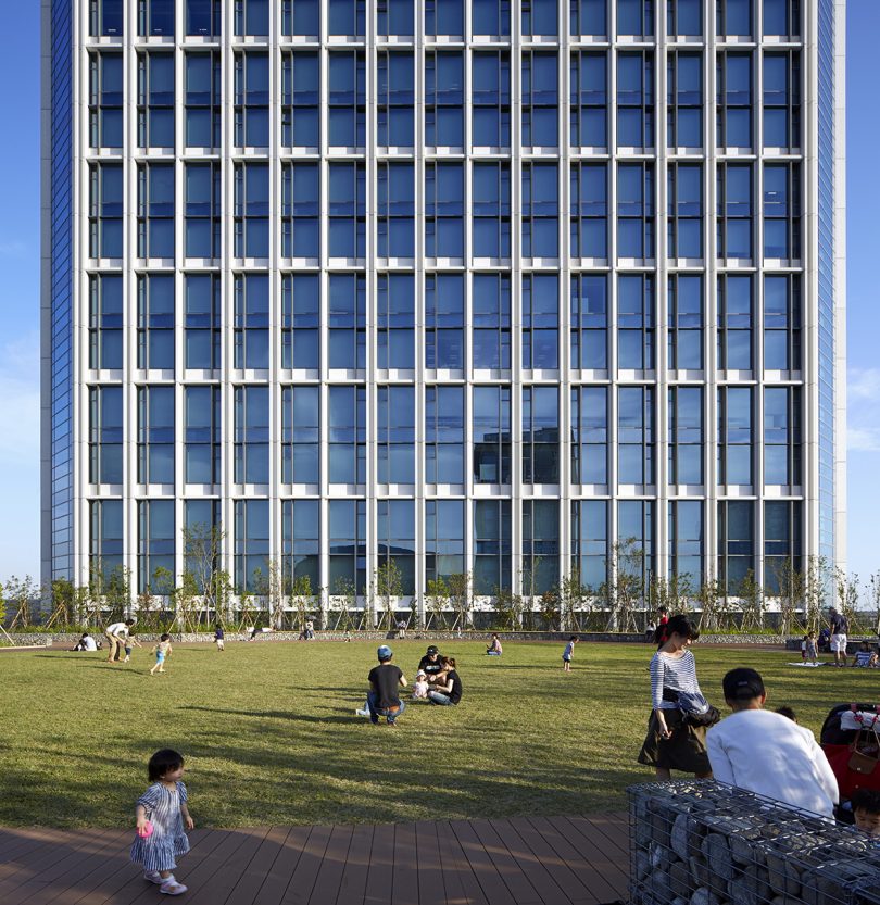 front facade of tall building with gridded windows, there are people relaxing in the grassy space in the foreground