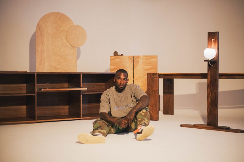 dark-skinned man sitting on the floor in front of a collection of wooden furniture