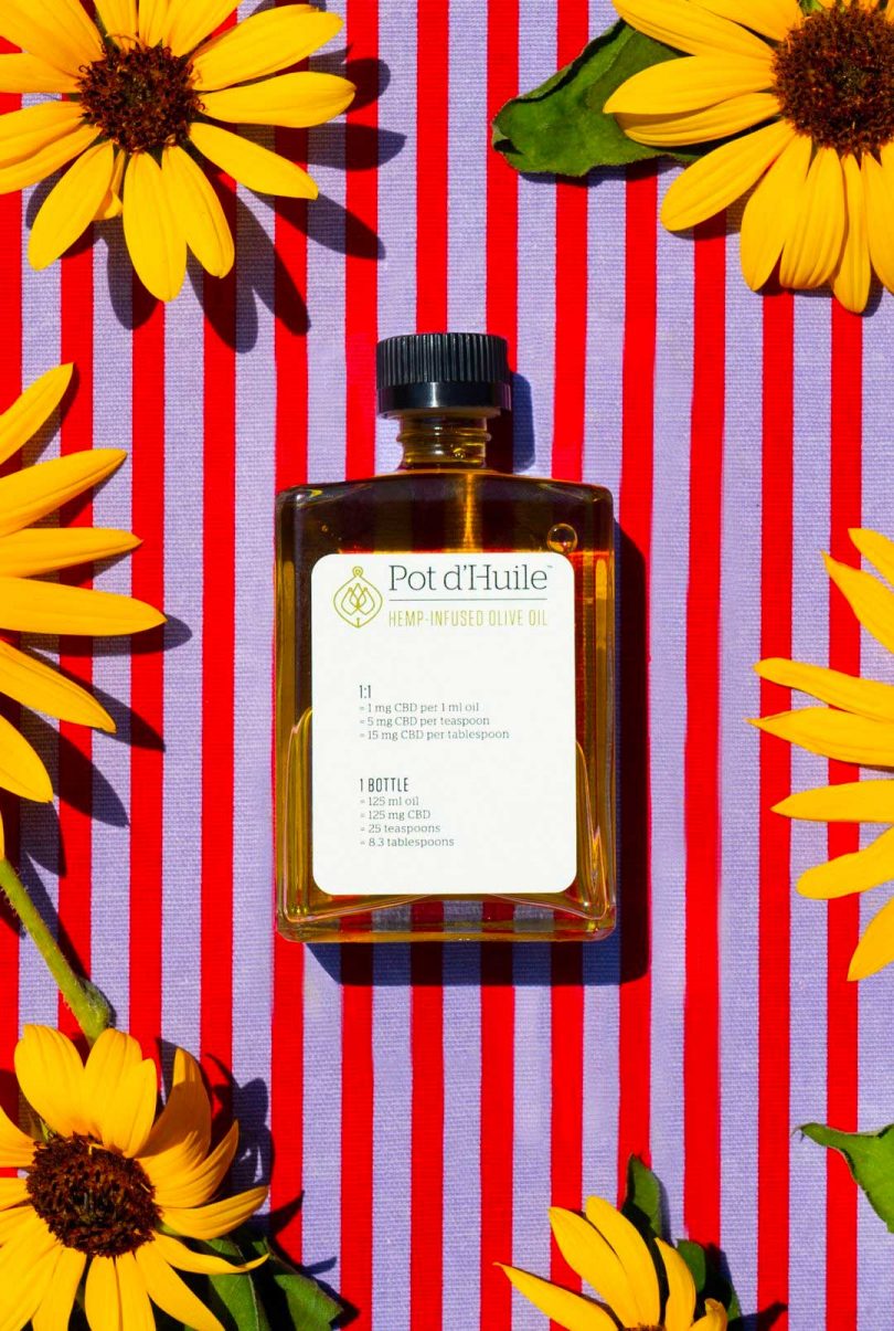 squared off bottle of brown liquid with white label with colorful background of red and lavender stripes and yellow sunflowers