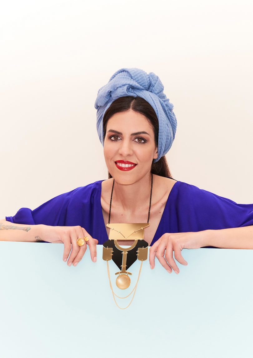 light-skinned woman with dark hair and a light blue head scarf wearings a royal blue top and modern jewelry