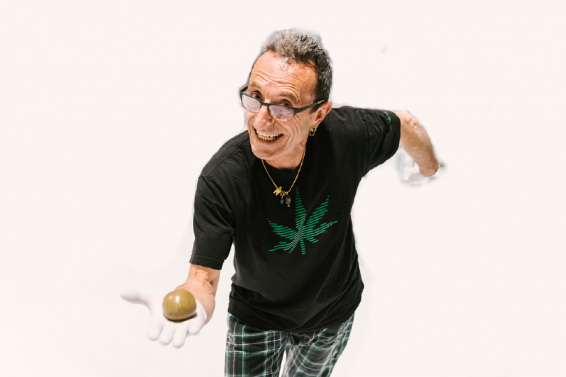 light-skinned man wearing glasses, a black shirt with a green cannabis leaf, and plain pants, holding out a pipe