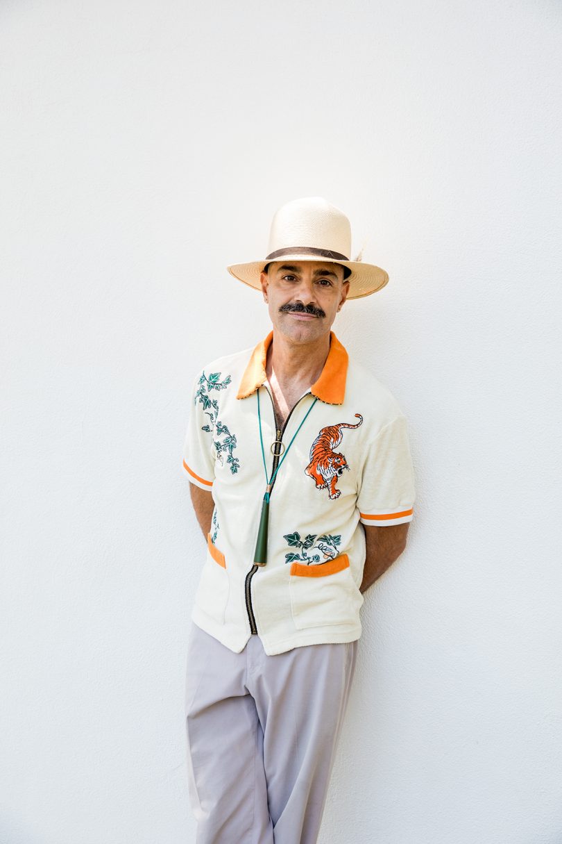light-skinned man with dark mustache wearing and orange and white collared button-up shirt and leaning against a white wall