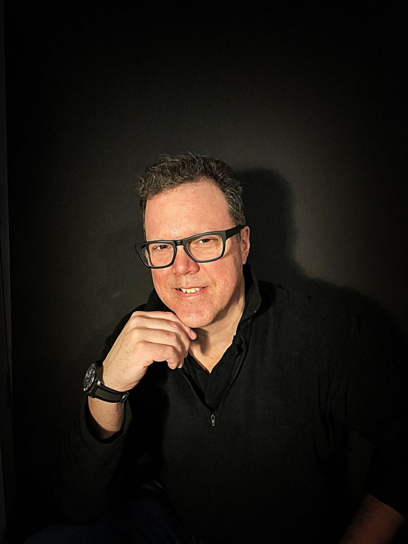 light-skinned man with dark hair and glasses wearing a black shirt and smiling at the camera with his chin resting on his right hand