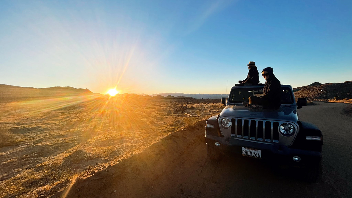 landscape vista of the sun rising or setting with two people watching from the hood of a Jeep