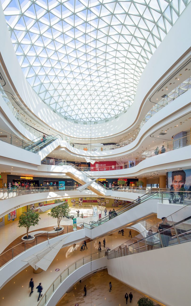تصویر عمودی از فضای داخلی یک فضای تجاری مدرن سفید با سطوح متعدد و سقف شیشه ای هندسی