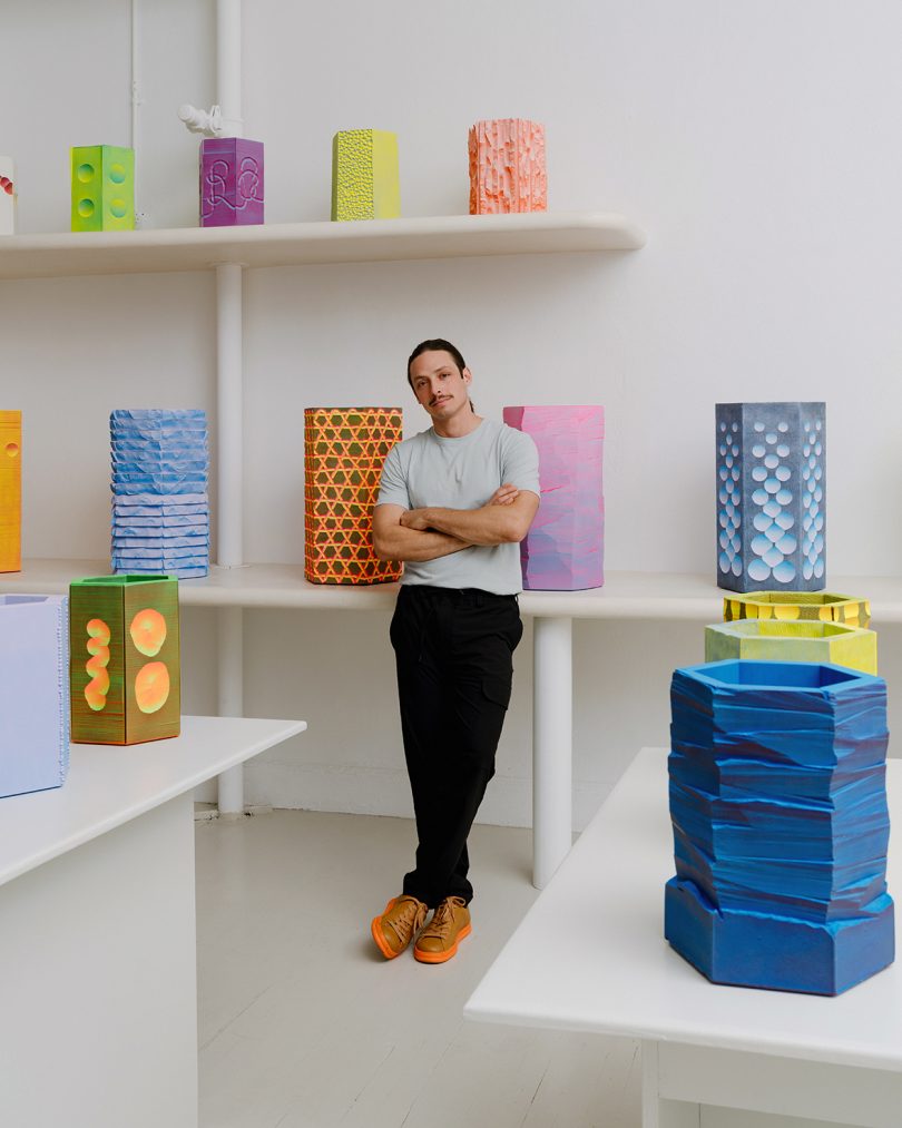 light-skinned man standing with his arms and legs crossed while leaning against a shelf in the middle of a white gallery space full of colorful vessels
