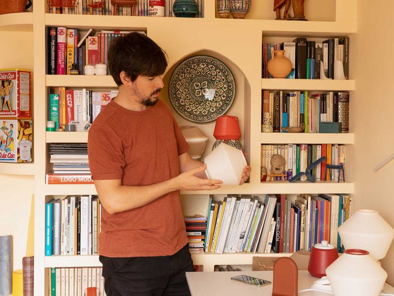 light-skinned man with dark hair and facial hair standing in front of loaded bookshelves admiring a lamp