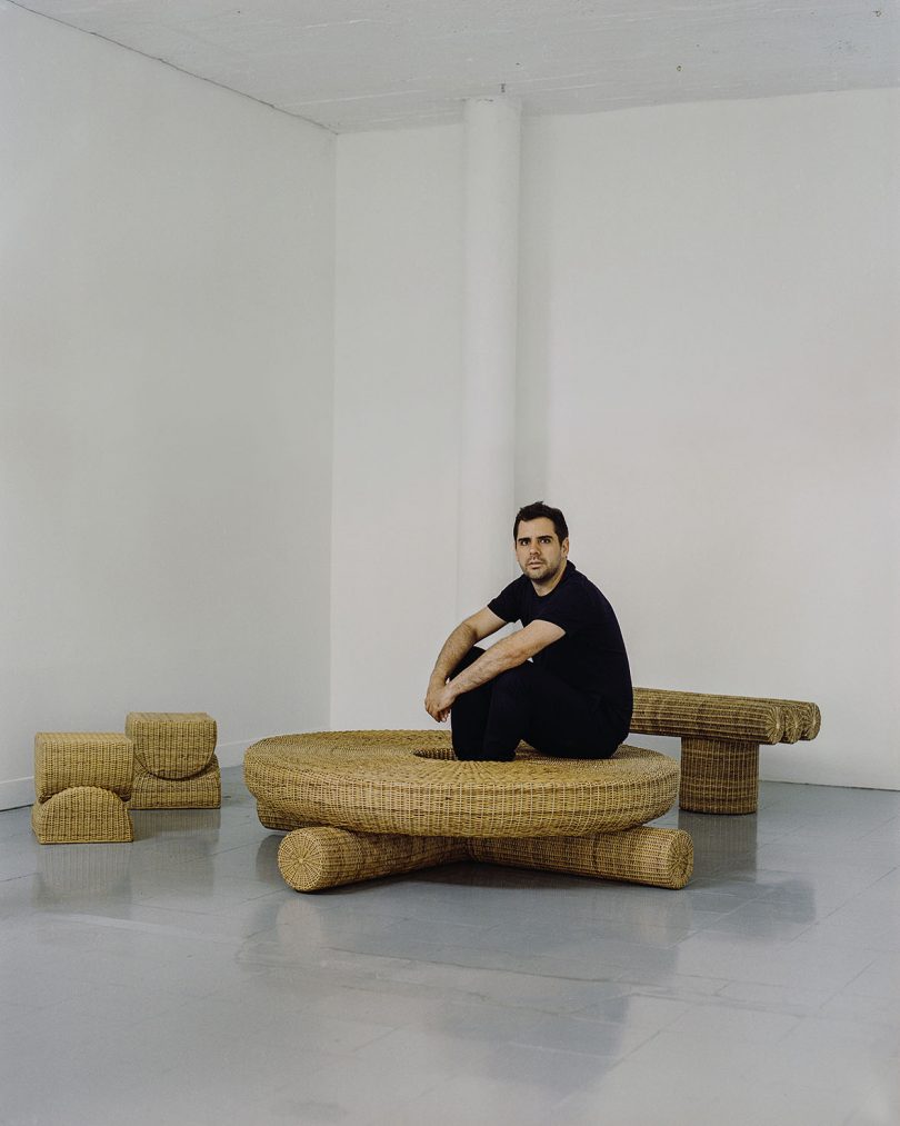 light-skinned man with dark hair, wearing black clothes sitting on a woven coffee table and surrounded by the rest of the collection