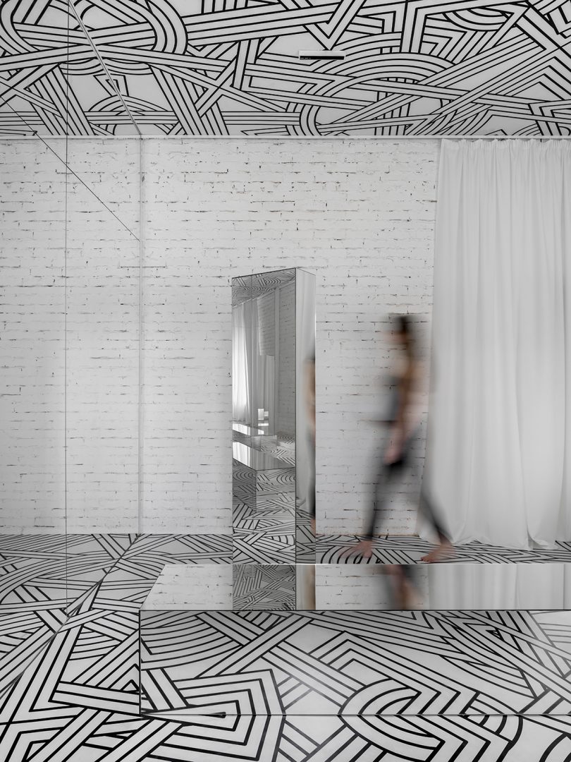 woman wearing all black walking in a small exhibition filled with a black and white geometric pattern and lots of mirrors