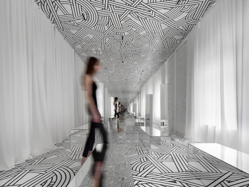 woman wearing all black walking in a small exhibition filled with a black and white geometric pattern and lots of mirrors