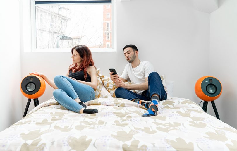 Orange OEPLAY pair on each side of a bed with man and woman posed enjoying audio.