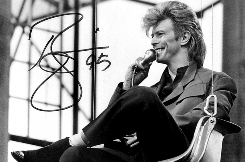 David Bowie sitting down, leaning against a wall, and smiling