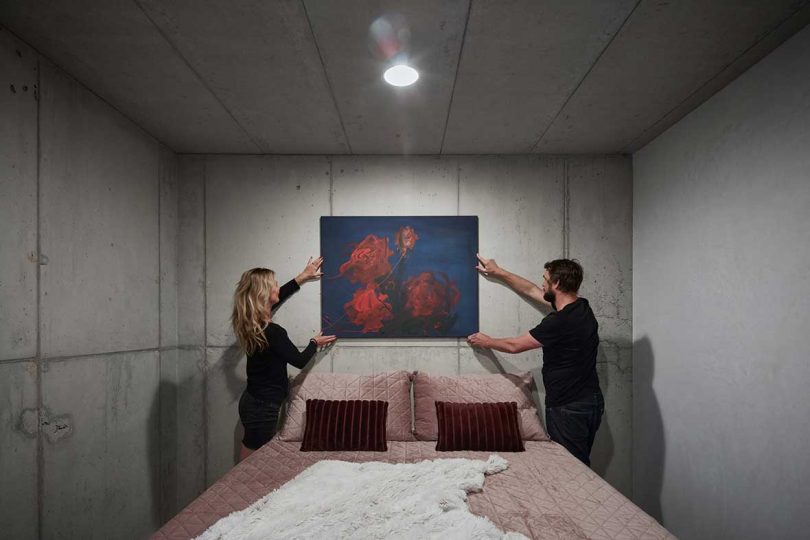 modern bedroom with concrete walls and two people hanging art over bed