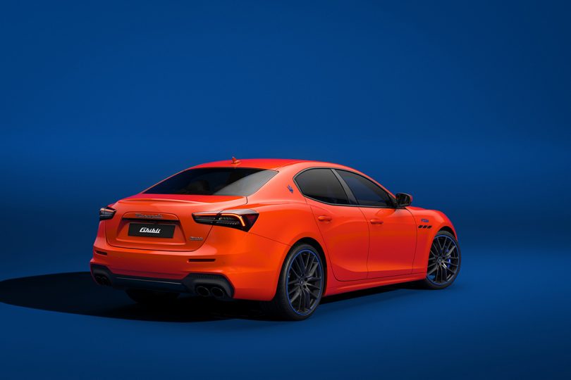 Rear view of Maserati Ghibli F Tributo in deep orange exterior paint against blue background.