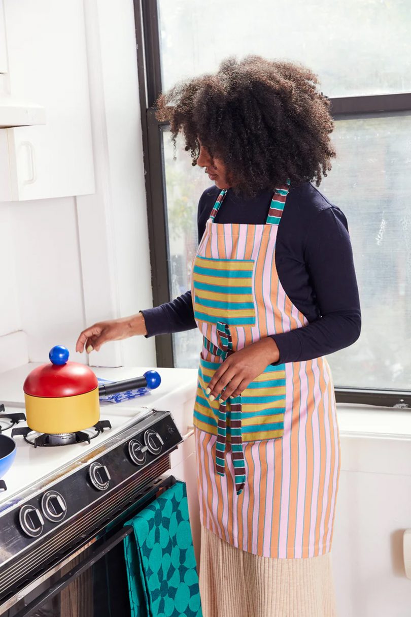 brown-skinned woman with dark curly hair cooking on a stove while wearing a green and pink striped apron