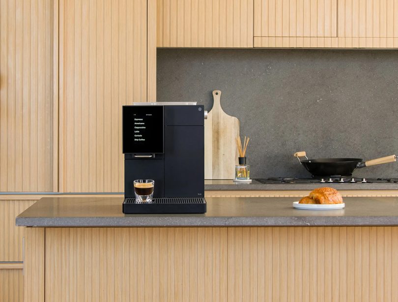 Front view of black TK-02 sitting on counter in modern kitchen with light honey wood slat cabinetry and a cutting board and wok in the background.