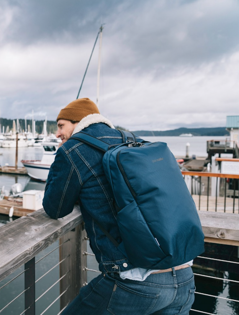 light-skinned man wearing a jean jacket and tan beanie with a blue backpack and leaning on a railing in a shipyard