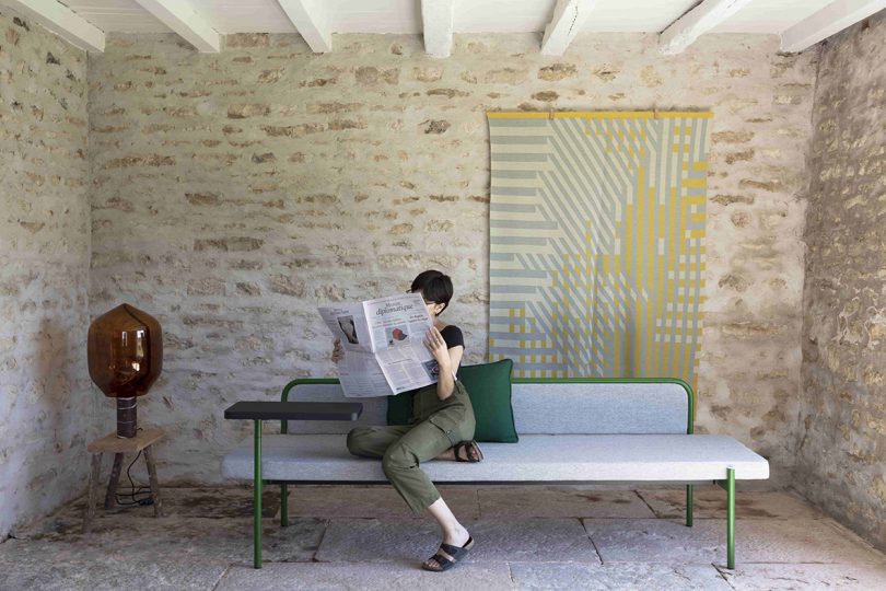 styled exterior space with sofa, end table, and a light-skinned woman reading the newspaper