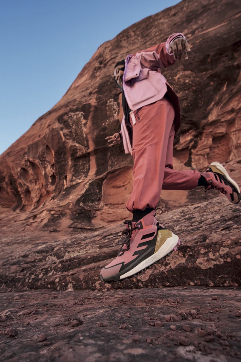 Angled ground level view of woman dressed in pink outdoor wear and in adidas Terrex Free Hiker 2 leaping-running across arid desert landscape. 