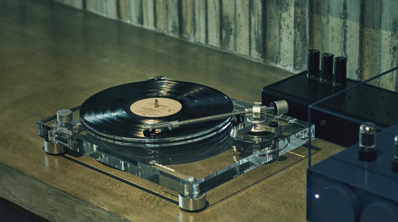 Audio-Technica 30 mm-thick, high-density clear acrylic chassis reduces resonance and reveals the belt-drive motor displayed on wood surface table with vacuum tube amps to the right.