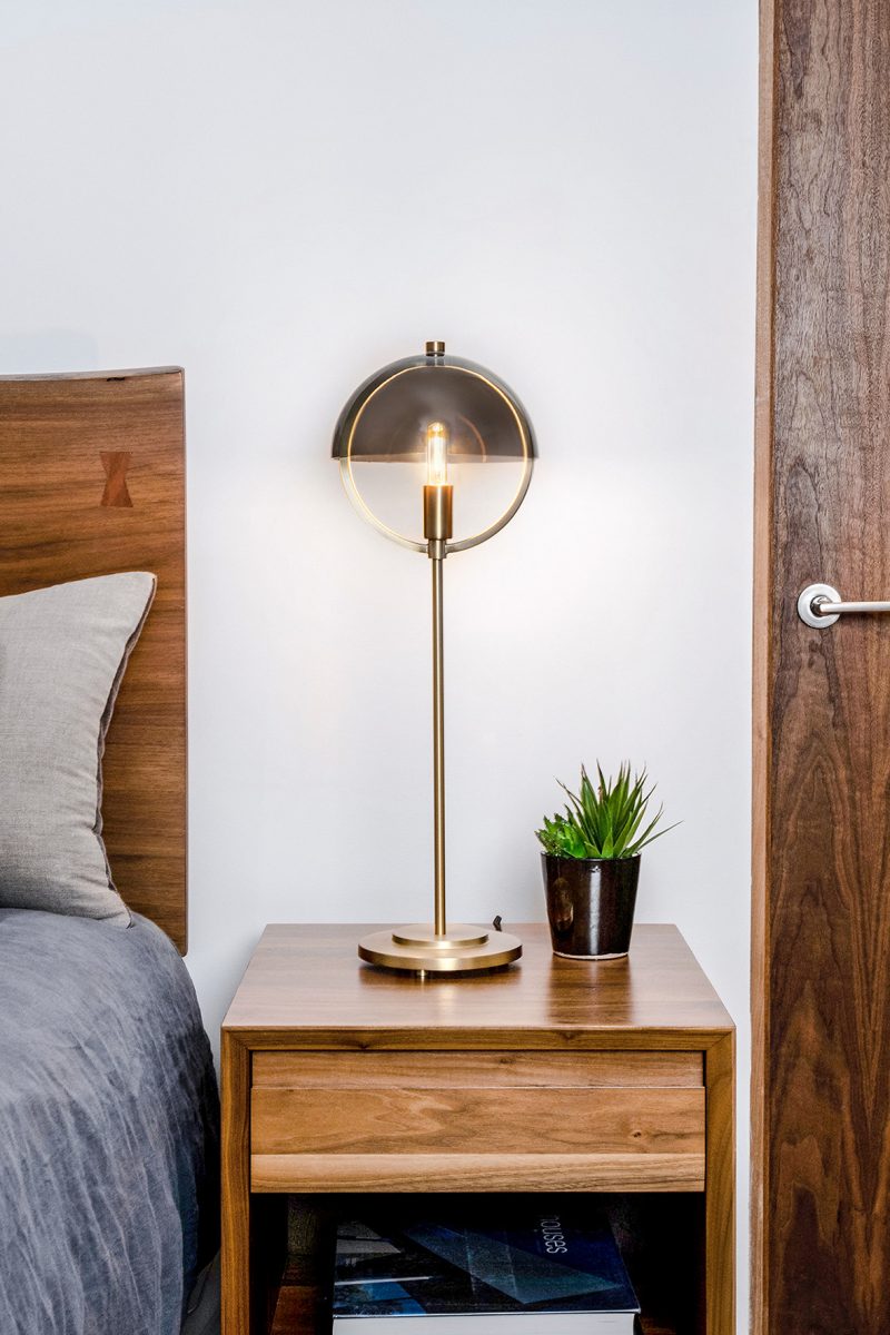 Table lamp version from the Copernica Collection set on nightstand in bedroom with wooden headboard and blue bedding.