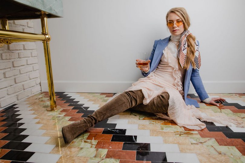 light-skinned blonde-haired woman wearing a dress and brown boots while sitting on a multi-color zigzag carpet