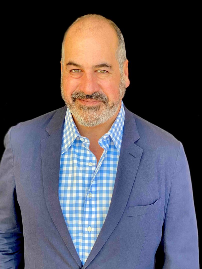 light-skinned man with grey hair and facial hair wearing a blue blazer and blue checked button-up shirt