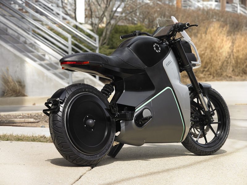 Rear angled view of FUELL Fllow electric motorcycle outdoors parked on sidewalk with stairs in background..