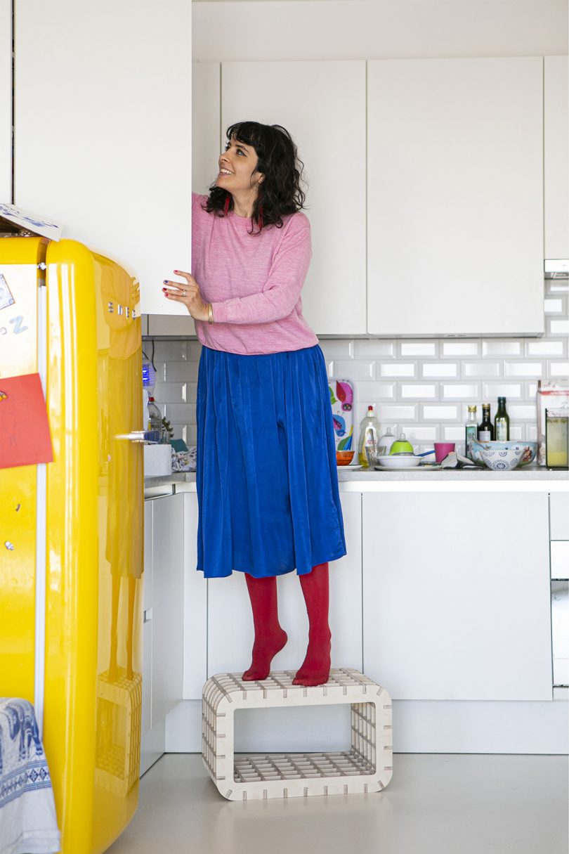 woman standing on a wood box-like stool to reach something in a kitchen cabinet