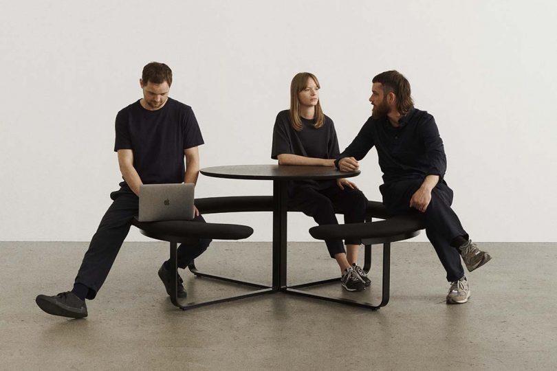 three people wearing black seated at a circular table surrounded by an attached circular bench