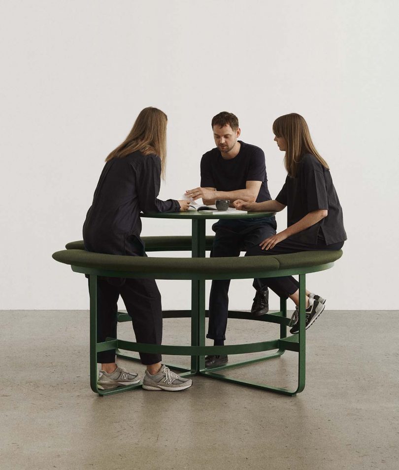 three people wearing black surrounding a circular table surrounded by an attached circular bench