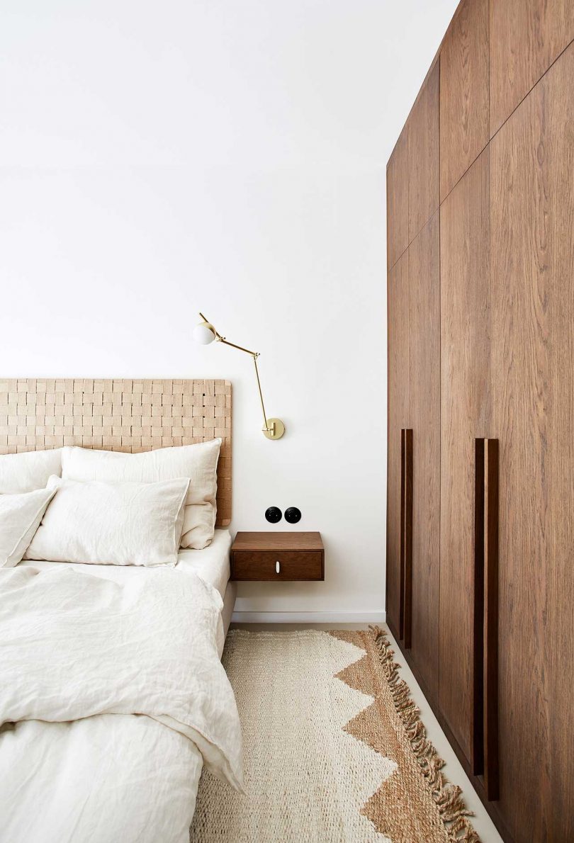 نیم نمای تخت خواب مدرن با تخت خواب روشن و روتختی کرم رنگ با دیوارهای کابینت چوبی در سمت راست