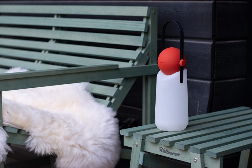 portable lamp with a white base, red top, and black strap handle sitting on a green wood outdoor side table