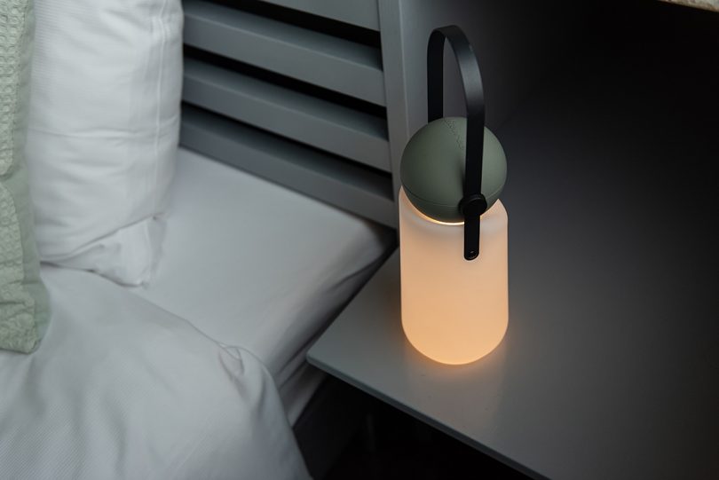 portable lamp with a white base, grey top, and black strap handle sitting on a bedside table