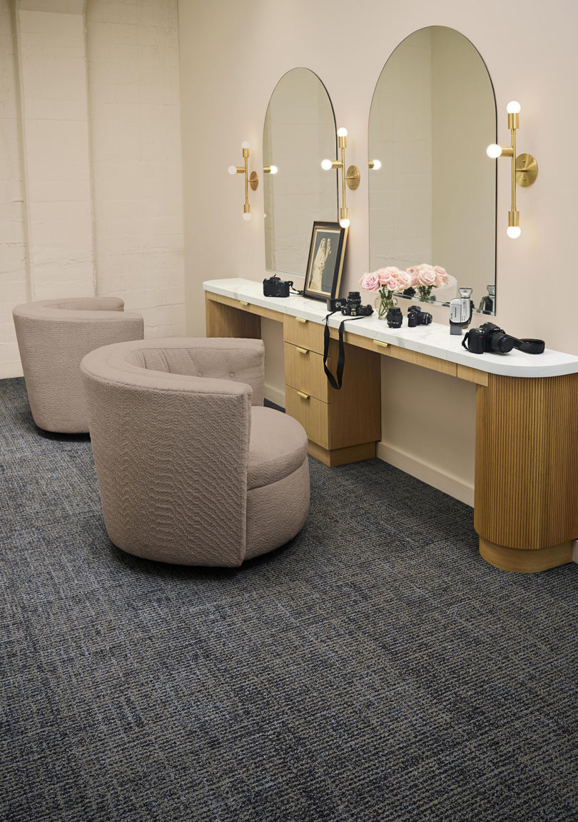 two wood and glass vanities side by side with round light pink armchairs and grey commercial carpeting