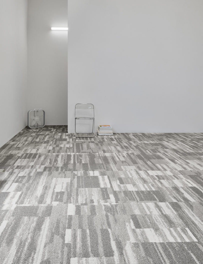 large space with geometric commercial carpeting in shades of grey, light grey walls, a folding chair, and a box fan