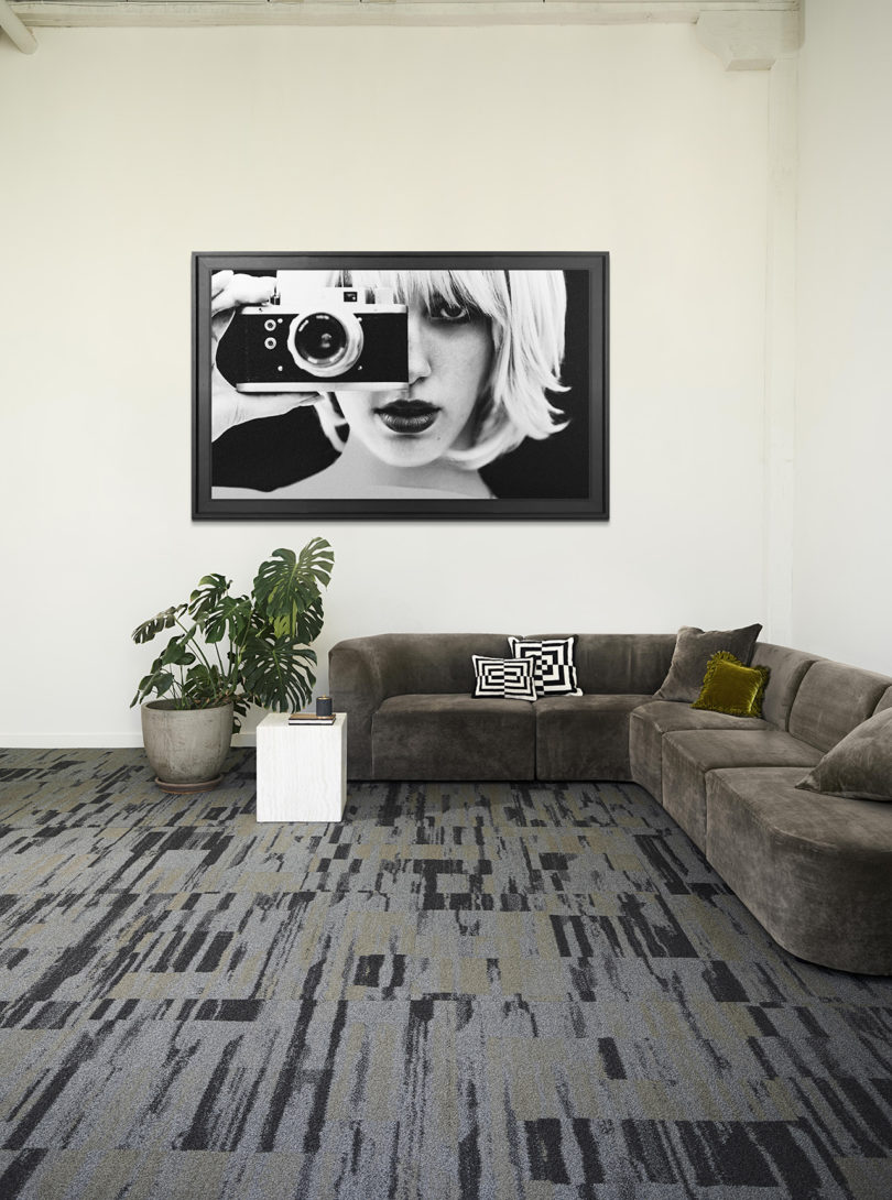 interior styled space with dark grey sectional sofa, white side table, potted floor plant, wall art, and dark grey patterned commercial carpeting