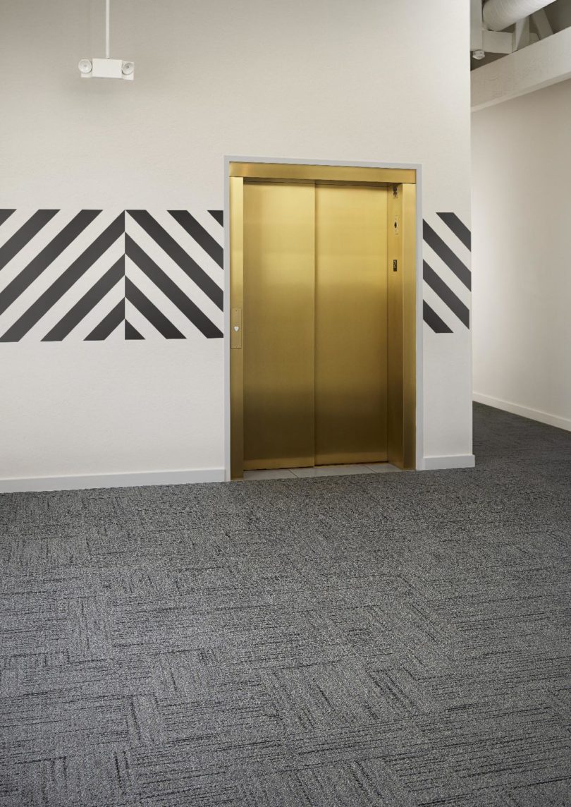 interior space with gold elevator doors, white walls with a banner of black and white pattern, and dark grey commercial carpeting