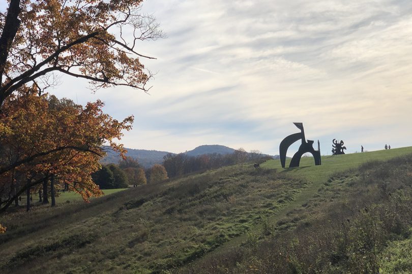 green field with colorful trees and an overcast sky over a large metal sculpture