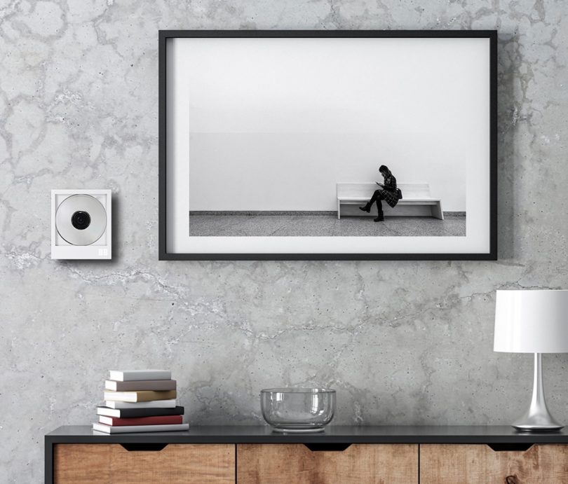 CP1 in living space setting, wall mounted above wood console with framed black and white photo to the right and small stack of books, clear bowl and small table lamp below.